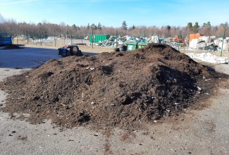 Esiplaanil on asfaldplatsil kuhi pruuni sõmera kompostiga, taaml näha prügikonteinerid ja -kuhjad